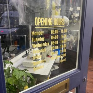 Business opening hours sign displayed on a shop window in West London, designed with gold lettering.